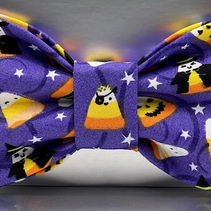May include: A purple Halloween-themed bow tie with a repeating pattern of candy corn, witch figures, and white stars. The bow tie is made of fabric and has a classic bow shape, perfect for festive occasions.