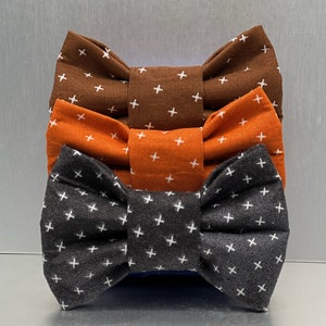May include: Three fabric bow ties stacked on top of each other, each with a cross pattern. The top bow tie is brown, the middle is orange, and the bottom is dark grey.