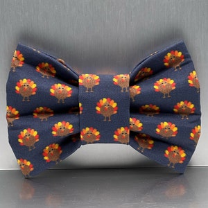 May include: A navy blue bow tie featuring a repeating pattern of small brown turkeys with yellow, orange, and red tail feathers. The fabric has a visible woven texture, and the bow tie is constructed with a central knot and two side wings.