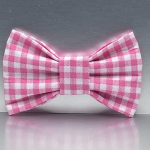 May include: A bow tie featuring a classic pink and white gingham check pattern. The fabric is folded into a structured, symmetrical shape with a central knot piece, displaying a consistent grid design across the entire surface of the accessory.