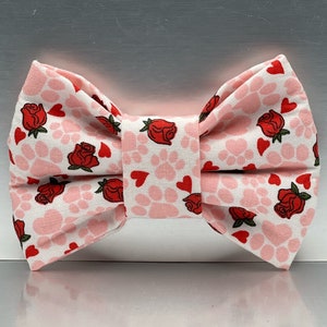 May include: A fabric bow tie featuring a repeating pattern of red roses, small red hearts, and light pink paw prints on a white background. The bow tie has a central fabric knot and is constructed with folded fabric to create a structured shape.