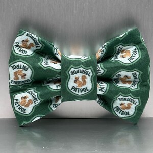 May include: A green bow tie featuring a repeating pattern of white shield-shaped patches. Each patch contains a brown squirrel illustration and the text SQUIRREL PATROL in black lettering. The fabric has a textured appearance throughout the design.