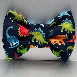 May include: A navy blue bow tie featuring a repeating pattern of dinosaurs in various colors including lime green, bright red, orange, and light blue. The fabric displays multiple species of dinosaurs with distinct shapes and spikes.