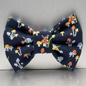 May include: A navy blue bow tie with a repeating pattern of colorful mushrooms. The mushrooms are red, yellow, and white, with green stems and leaves. The bow tie is made of fabric.
