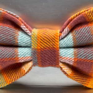 May include: A tartan bow tie in shades of orange, teal, and burgundy. The fabric has a textured appearance, with a classic plaid pattern. The bow tie is knotted in the centre and ready to wear.