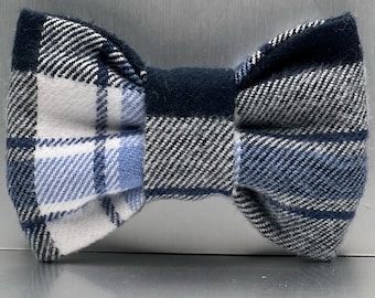 May include: A bow tie featuring a textured wool-like fabric with a plaid pattern in shades of navy blue, light blue, grey, and white. The design includes a solid navy blue center knot and a classic bow shape with distinct folded fabric edges.