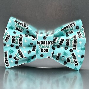 May include: A light blue bow tie featuring a repeating pattern of black paw prints, teal starbursts, and the text "WORLD'S BEST DOG" printed in black and teal lettering across the fabric surface in a structured, symmetrical arrangement.