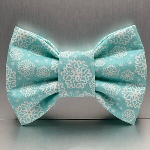 May include: A turquoise bow tie featuring a white snowflake pattern. The fabric bow tie has a classic shape. The snowflake design is evenly distributed across the fabric, creating a festive look.