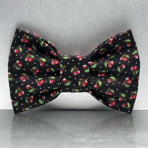 May include: A black bow tie featuring a repeating pattern of small red cherries with green stems and leaves, interspersed with tiny white polka dots across the entire fabric surface of the accessory, which is shaped in a classic butterfly style.