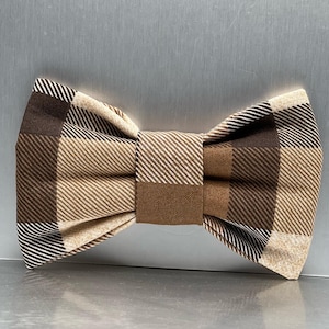 May include: A brown and beige checked bow tie. The bow tie has a classic plaid design in shades of brown and beige. The fabric is folded and sewn into a bow shape, with a central knot and symmetrical wings. The bow tie is designed to be worn around the neck.