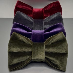 May include: A set of four velvet bow ties arranged in a vertical stack. The colors from top to bottom are deep red, charcoal gray, royal purple, and olive green. Each bow tie features a central fabric knot and a soft, textured velvet finish.