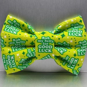May include: A bright lime green bow tie featuring a repeating white pattern of four-pointed stars and the text "Rub my belly for GOOD LUCK" printed in a bold, sans-serif font across the fabric surface in a consistent, repeating layout.