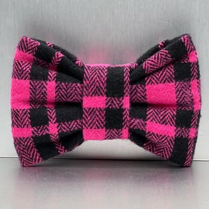 May include: A pink and black plaid bow tie. The bow tie is made of a soft, fuzzy fabric.