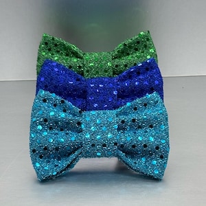 May include: A stack of three bow ties featuring a textured, glittery finish. The top bow tie is green, the middle bow tie is royal blue, and the bottom bow tie is a bright turquoise blue. Each bow tie has a classic shape with a central knot.