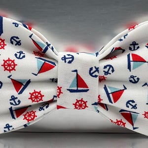 May include: A white bow tie with a nautical pattern featuring red and blue sailboats, anchors, and ship wheels. The bow tie is a classic accessory for formal or casual wear, adding a touch of maritime flair.