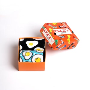 May include: A set of two pairs of socks in a bright orange box. The socks feature a fun, colourful design of fried eggs on a black and blue background. The box has the text "OZZY SOCKS" printed on it.