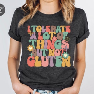 May include: Heather dark grey t-shirt with a retro-style graphic that says "I tolerate a lot of things but not gluten". The graphic features colorful letters, flowers, and smiley faces.