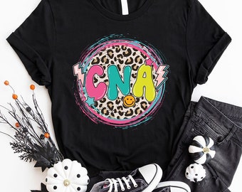 May include: Black t-shirt with a colorful graphic of the word "CNA" in a circle with a leopard print background. The graphic also includes a smiley face and lightning bolts.