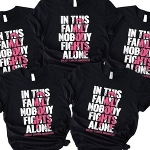 May include: Five black t-shirts with a pink ribbon graphic and the text "In this family nobody fights alone" printed on the front. The text is in a distressed font style. The shirts are a symbol of breast cancer awareness.