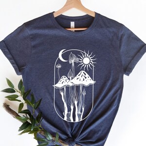 May include: Navy blue t-shirt featuring a white graphic of mushrooms, a crescent moon, and a sun within an oval frame. The shirt is made of a soft material and has rolled-up sleeves. The design is a nature-inspired illustration.