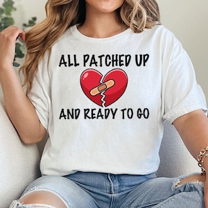 May include: White t-shirt with a red heart graphic with a bandage on it. The text on the shirt reads "All patched up and ready to go."