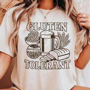 May include: A cream-colored t-shirt featuring a dark brown graphic print with the text "GLUTEN TOLERANT" and illustrations of a bowl of pasta, a mug of beer, wheat stalks, and a loaf of sliced bread arranged in a central design on the front.