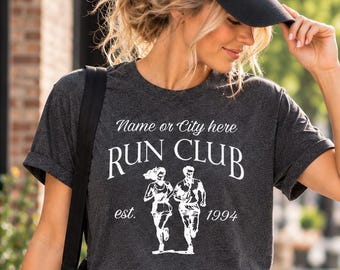 May include: A dark charcoal gray short-sleeve t-shirt featuring white text that reads Name or City here RUN CLUB est. 1994, accompanied by a graphic illustration of two people running. The shirt is paired with black athletic shorts and a black cap.