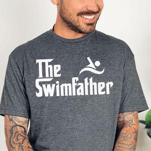 May include: Dark gray t-shirt with the text "The Swimfather" in white, featuring a swimmer graphic. The shirt is made of a soft material and is suitable for casual wear. The person in the image is wearing blue jeans.