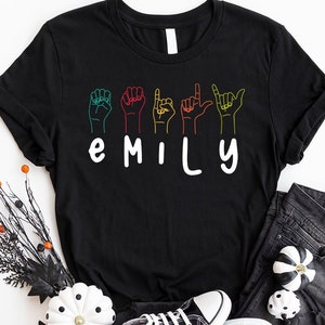 May include: Black t-shirt with the name "EMILY" spelled out in white letters. Each letter is represented by a different colored hand sign in American Sign Language.