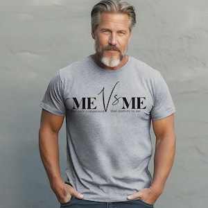 May include: A heather gray short-sleeved t-shirt featuring the black text ME VS ME centered on the chest. Below the main text, smaller black lettering reads The only comparison that matters to me. The shirt has a crew neckline and a standard fit.