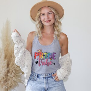 May include: A heather gray racerback tank top featuring the text "PRIDE tribe" in a colorful, rainbow-gradient font. The design includes a small crown above the word "PRIDE" and three black hearts. The tank top is worn with a white open-knit cardigan.
