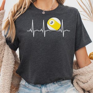 May include: A dark heather gray short-sleeve t-shirt featuring a white EKG heartbeat line graphic across the chest. Integrated into the heartbeat line is a yellow and white billiard ball with the number nine printed in black on its surface.