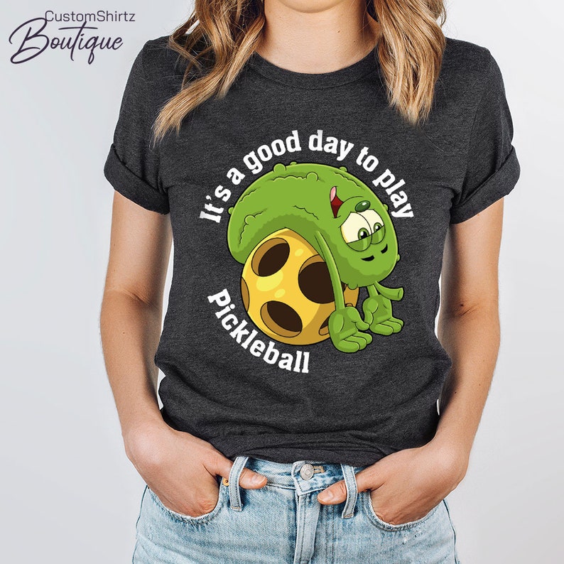 May include: A dark gray t-shirt with a green pickle character holding a yellow pickleball. The text "It's a good day to play Pickleball" is printed on the shirt.