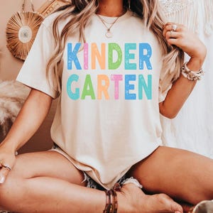 May include: A cream-colored short-sleeve t-shirt featuring the word KINDERGARTEN printed in large, distressed, multi-colored block letters. The text is arranged in two rows with the letters in shades of blue, orange, green, and pink.
