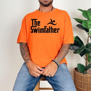 May include: Bright orange t-shirt with the text "The Swimfather" in black. The design includes a swimmer icon. The shirt is a casual style, suitable for everyday wear.