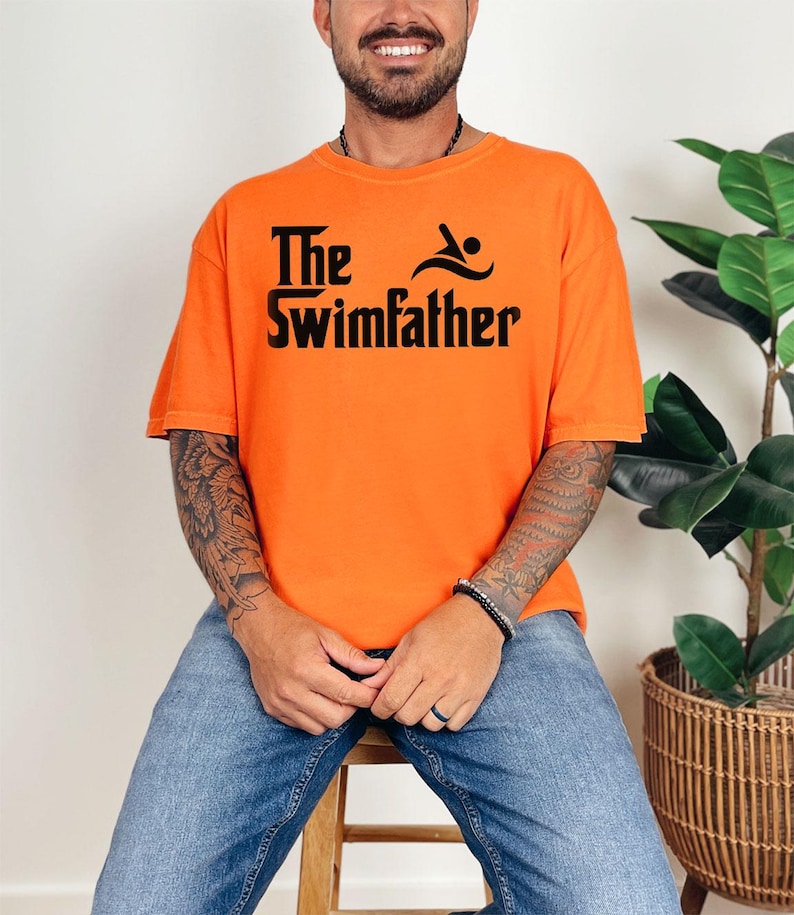May include: Bright orange t-shirt with the text "The Swimfather" in black. The design includes a swimmer icon. The shirt is a casual style, suitable for everyday wear.
