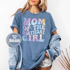 A woman wearing a blue t-shirt with the text "MOM OF THE BIRTHDAY GIRL" printed on it, along with a denim jacket and denim shorts.