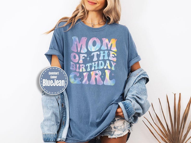 A woman wearing a blue t-shirt with the text "MOM OF THE BIRTHDAY GIRL" printed on it, along with a denim jacket and denim shorts.