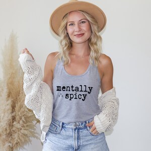 May include: A light gray tank top featuring the black text 'mentally spicy' with small star icons. The garment is worn with a cream-colored open-knit cardigan, a tan wide-brimmed hat, and light blue denim shorts, creating a casual layered look.