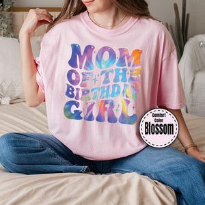 A woman wearing a pink t-shirt with the text "MOM OF THE BIRTHDAY GIRL" is sitting on a bed.