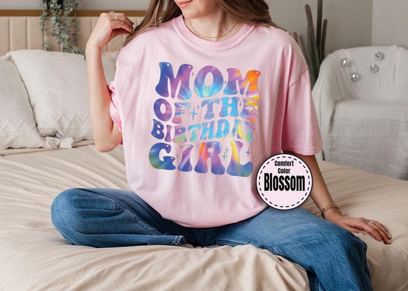 A woman wearing a pink t-shirt with the text "MOM OF THE BIRTHDAY GIRL" is sitting on a bed.