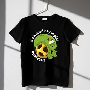 May include: Black t-shirt with a green cartoon pickle character holding a yellow pickleball. The text "It's a good day to play pickleball" is printed on the shirt.