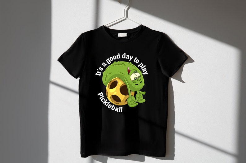 May include: Black t-shirt with a green cartoon pickle character holding a yellow pickleball. The text "It's a good day to play pickleball" is printed on the shirt.