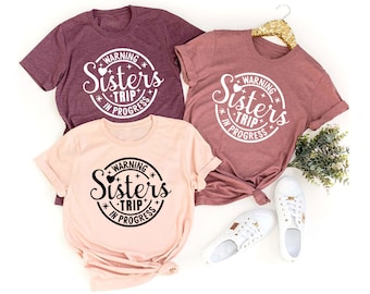 May include: Three t-shirts with a black and white graphic design that says "Warning Sisters Trip in Progress". The shirts are in different colors: maroon, pink, and light pink.