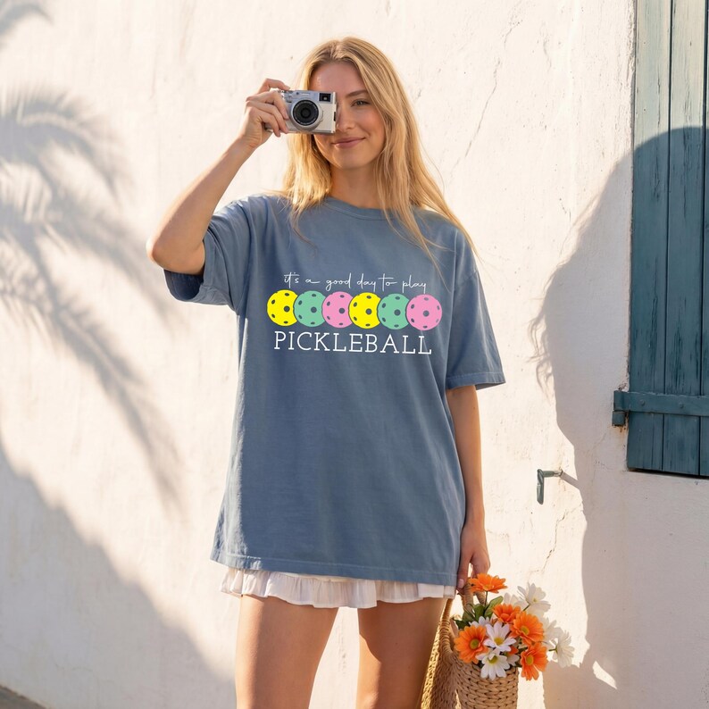 A woman wearing a blue t-shirt with a graphic of a pickleball and holding a basket of flowers, smiling and holding a camera up to her face.