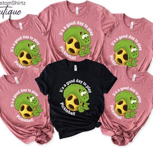 May include: Five pink and black t-shirts with a green pickleball graphic and the text "It's a good day to play Pickleball".
