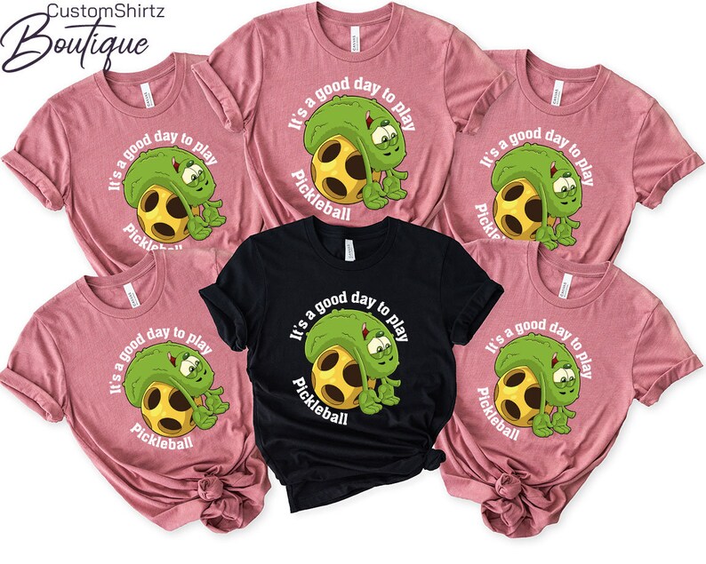 May include: Five pink and black t-shirts with a green pickleball graphic and the text "It's a good day to play Pickleball".