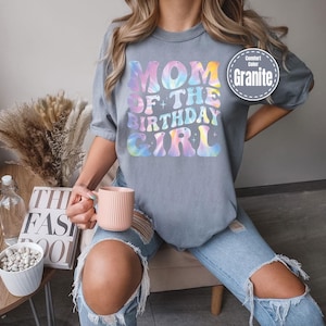 A person wearing a gray t-shirt with the text "MOM OF THE BIRTHDAY GIRL" printed on it, holding a pink mug and sitting on a chair.