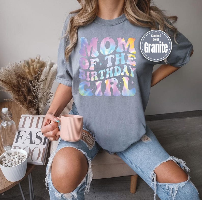 A person wearing a gray t-shirt with the text "MOM OF THE BIRTHDAY GIRL" printed on it, holding a pink mug and sitting on a chair.