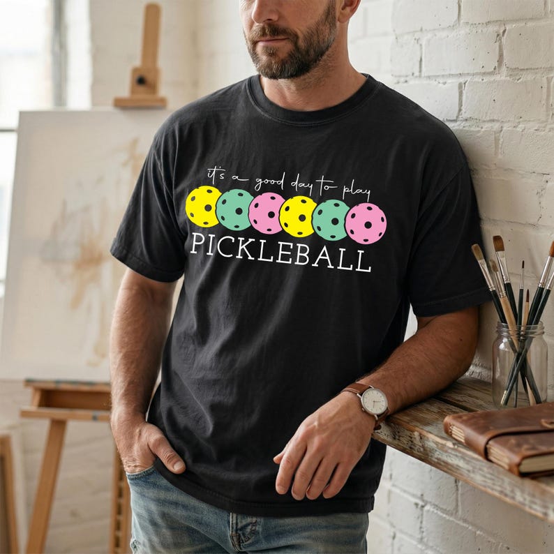 A man wearing a black t-shirt with a graphic design of colorful pum balls and the text "It's a good day to play PICKLEBALL" is standing in front of a wooden shelf with art supplies.